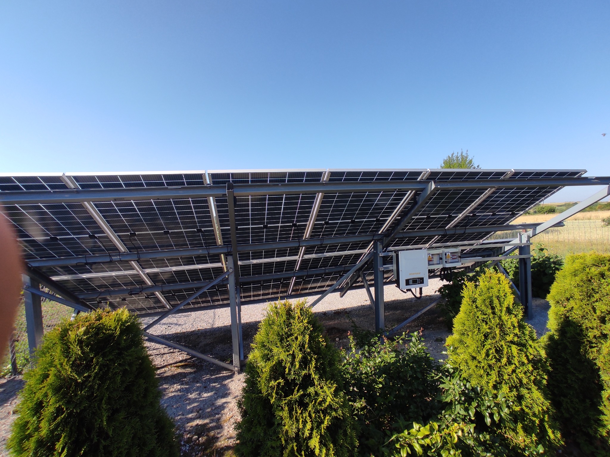 Widok od dołu na panele fotowoltaiczne zamontowane na metalowej konstrukcji, z widocznym inwerterem i zielenią w tle, w słoneczny dzień.