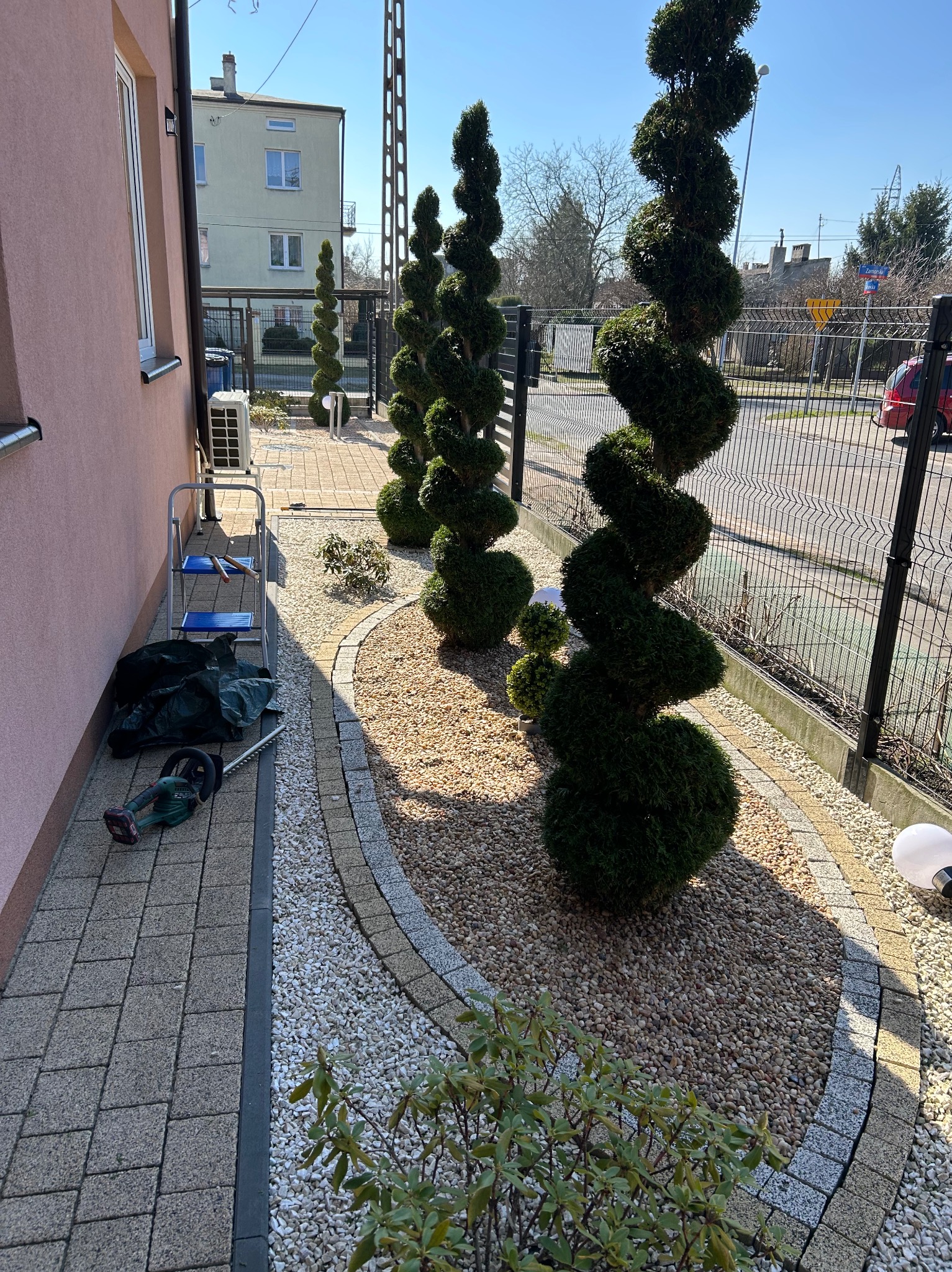 Ogród z formowanymi spiralnie tujami, ścieżką z kamyków i kostki brukowej. W tle budynek mieszkalny i narzędzia ogrodnicze.