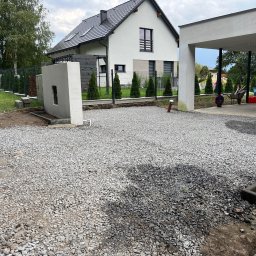 JB Nawierzchnie SP. Z o.o. - Przygotowany teren pod budowę z wysypanym żwirem, widok na nowoczesny dom z szarą dachówką i ogrodzeniem z tujami. Częściowo zadaszone miejsce parkingowe.