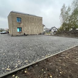 JB Nawierzchnie SP. Z o.o. - Utwardzony teren żwirem przy nowoczesnym domu z płaskim dachem, obrzeża z kostki. Widoczne drzewa i sąsiednie budynki w tle, pochmurne niebo.