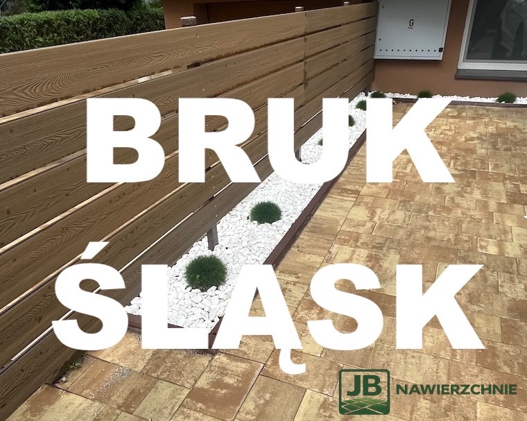 Ułożona kostka brukowa w odcieniach beżu, drewniane ogrodzenie i rabata z białym kamieniem oraz zielonymi kulami. Widoczny fragment elewacji budynku.