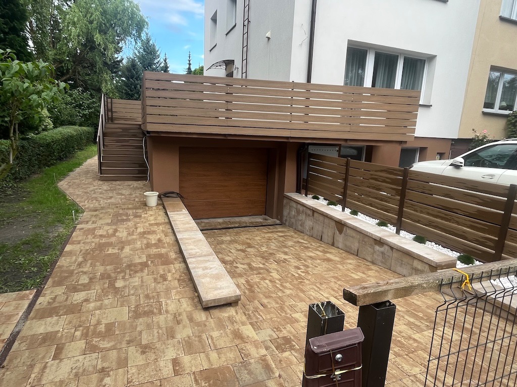Ułożona kostka brukowa w odcieniach brązu prowadząca do garażu z drewnianą bramą. Widoczne schody, ogrodzenie z drewnianych paneli i fragment białego samochodu.