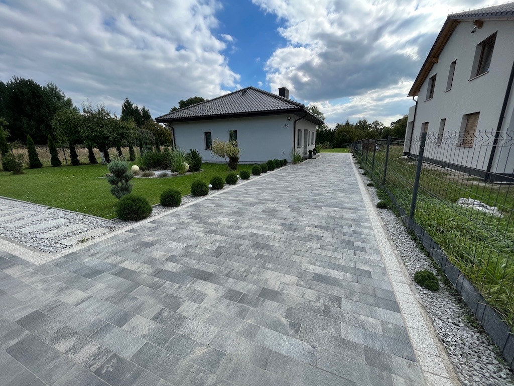 Szara kostka brukowa prowadząca do domu z numerem 39, obok trawnik z ozdobnymi krzewami i ogrodzenie. Widok na posesję w słoneczny dzień.