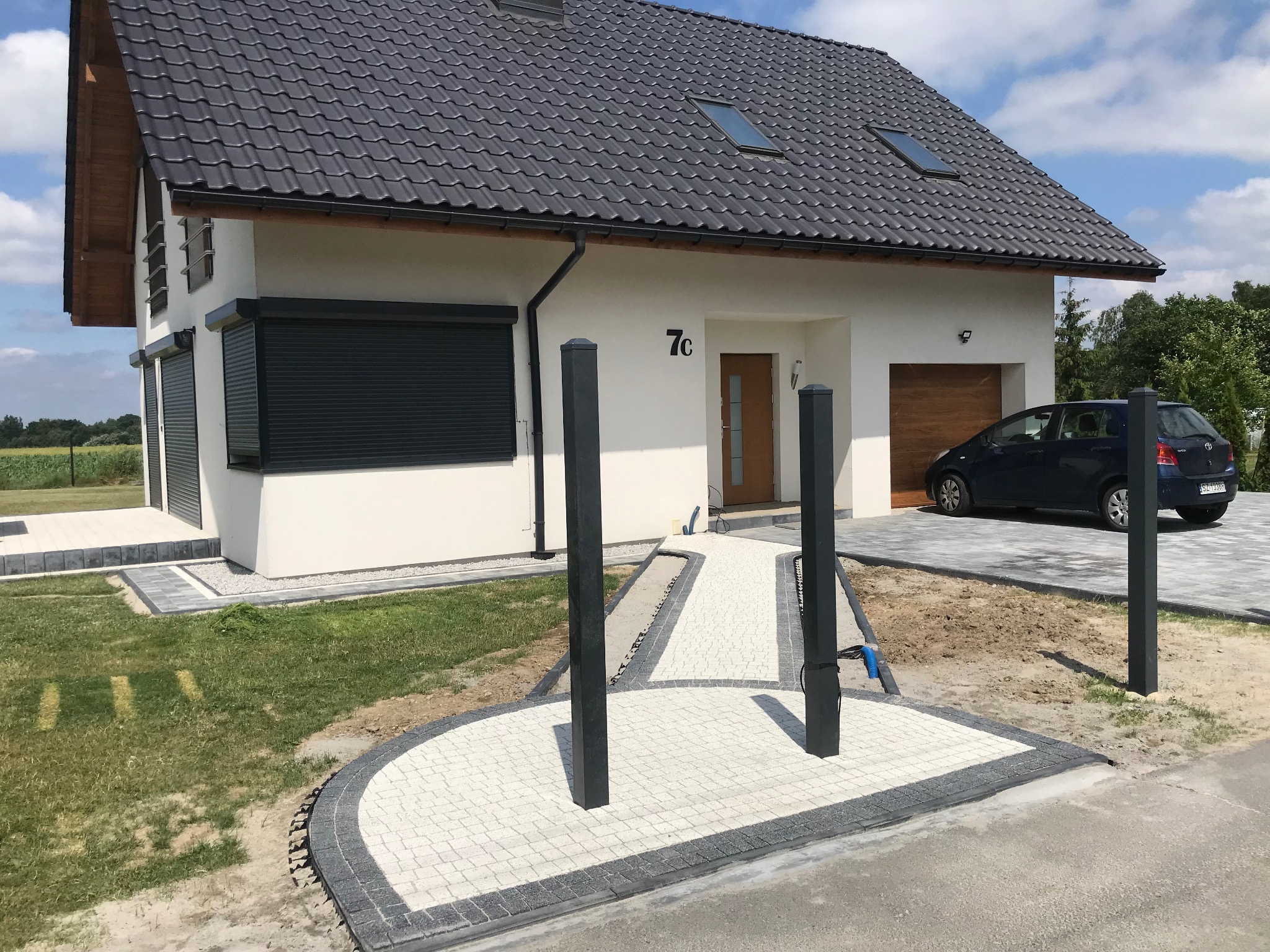 Nowoczesny dom z jasną kostką brukową tworzącą ścieżkę i okrągły podjazd. Ciemna obwódka kostki, słupki oświetleniowe. W tle dom z garażem i samochód.