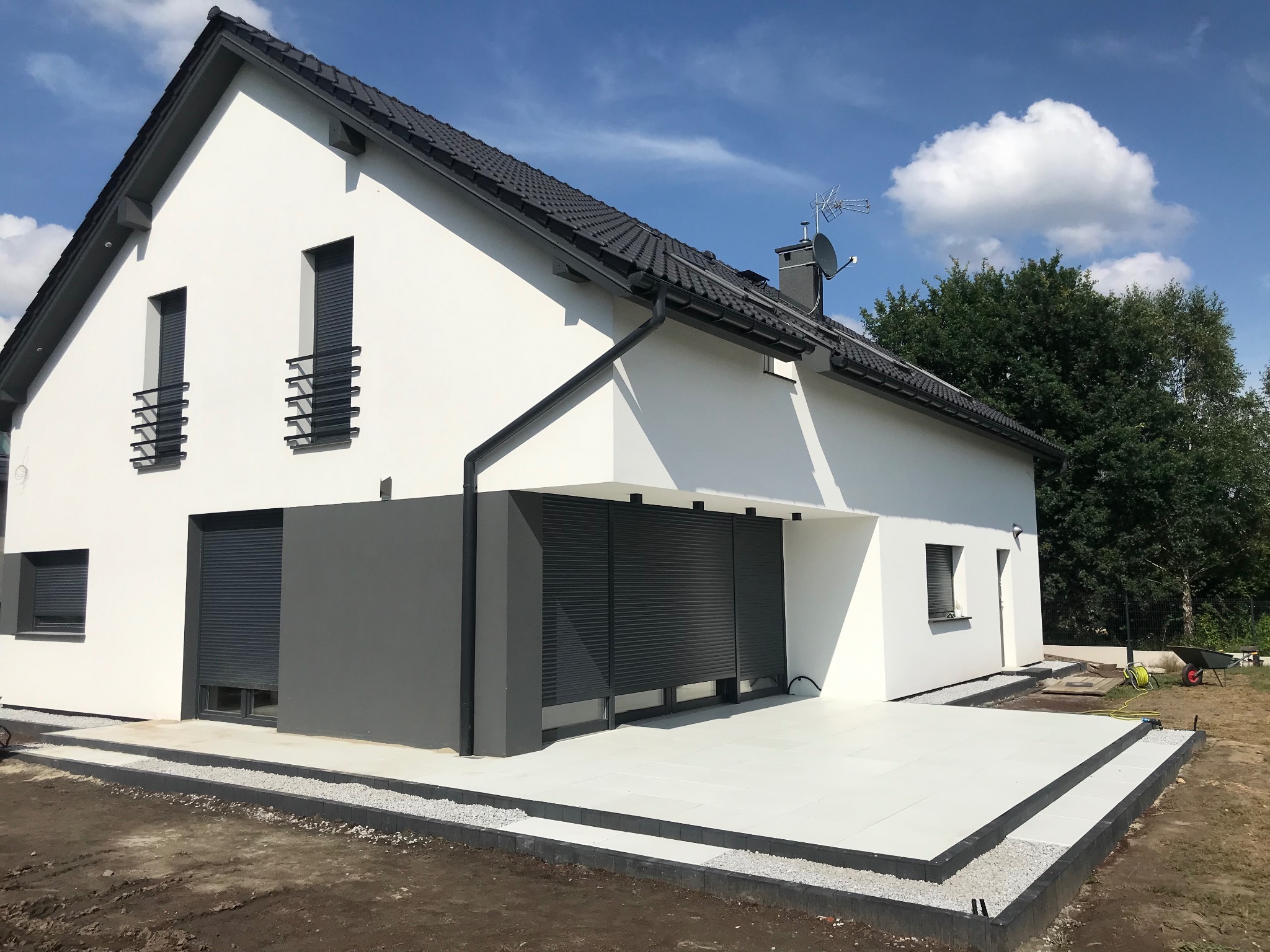 Nowoczesny dom z tarasem wyłożonym jasnymi płytami. Widoczne obrzeża z ciemnej kostki i jasny żwir. Elewacja biała z szarymi roletami. Błękitne niebo z chmurami w tle.