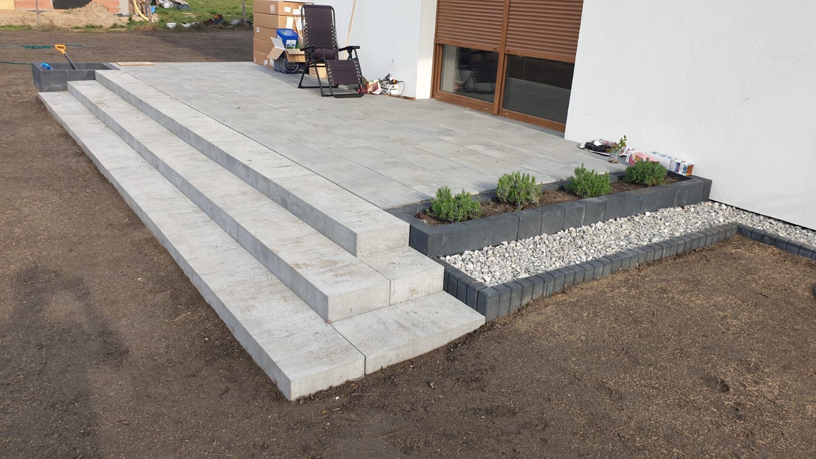 Nowoczesny taras z jasnoszarej kostki brukowej, schody z bloków betonowych, obrzeża z ciemnej kostki, rabata z kamykami i roślinami. Widok na dom z roletami.