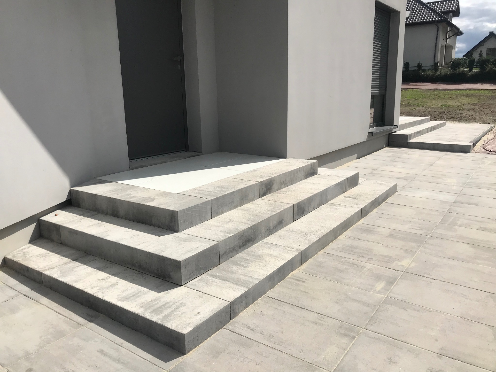 Nowoczesne schody wejściowe z szarej kostki brukowej prowadzące do domu z szarą stolarką okienną i drzwiową. Estetyczne wykończenie terenu.