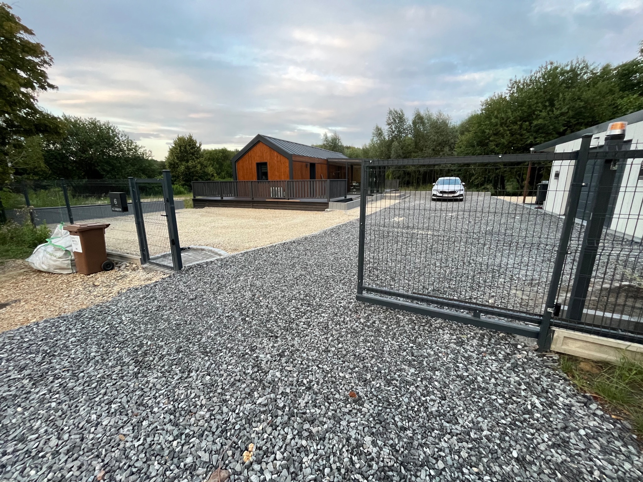 Żwirowa droga do nowoczesnego domu z drewnianą elewacją, widoczna przez otwartą, metalową bramę. W tle zaparkowany samochód i zieleń drzew.