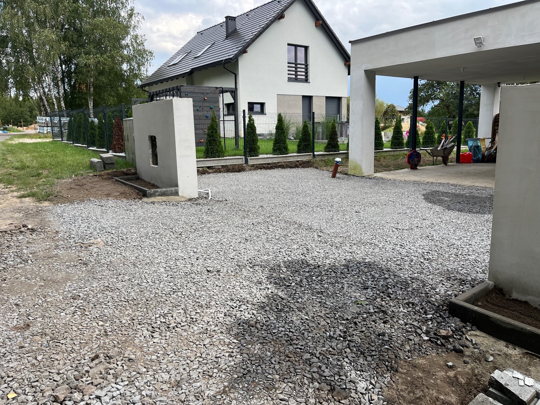 Przygotowany teren pod budowę z wysypanym żwirem, widok na nowoczesny dom z szarą dachówką i ogrodzeniem z tujami. Częściowo zadaszone miejsce parkingowe.