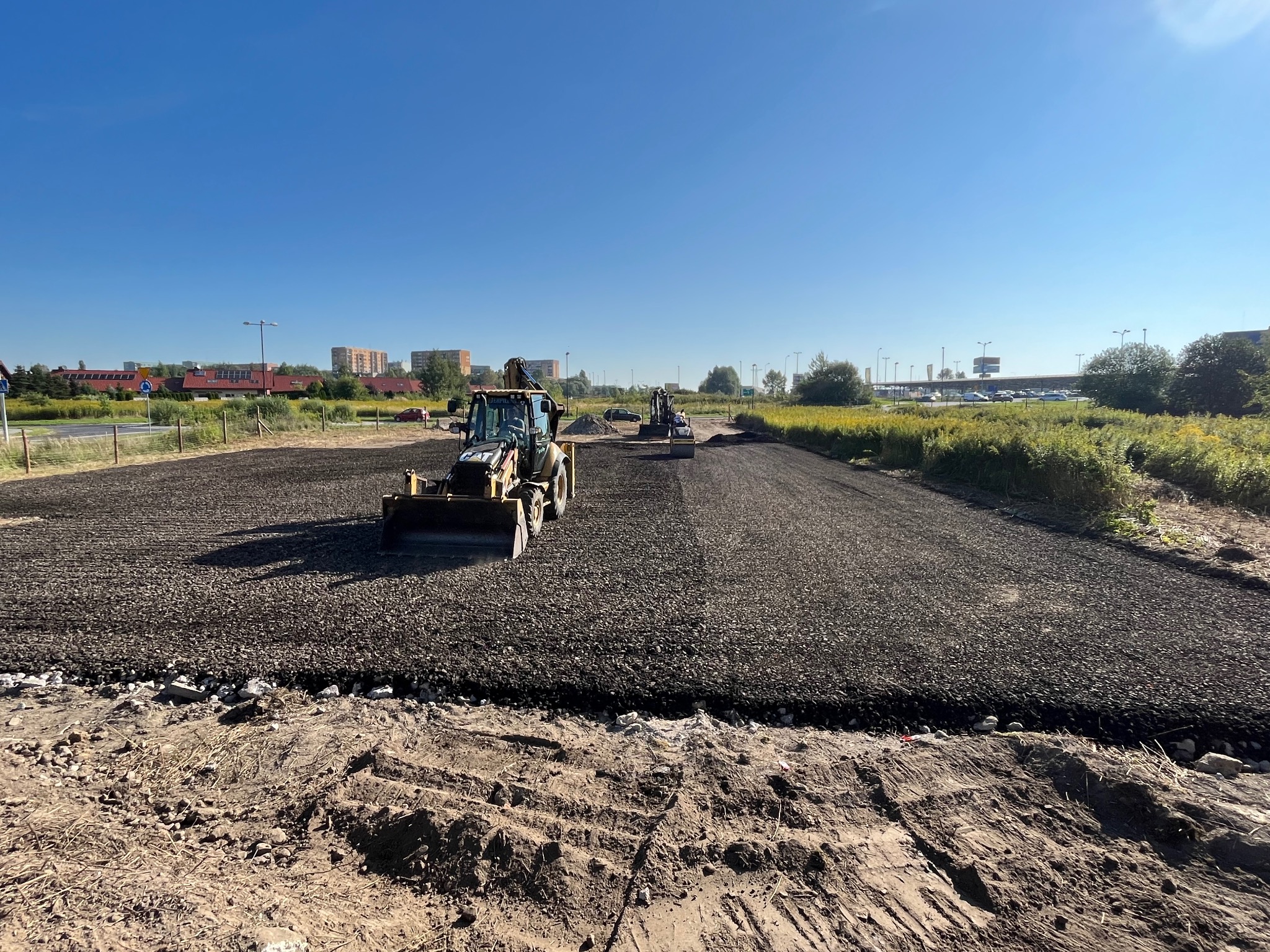Żółty traktor równa powierzchnię drogi pokrytej drobnym kruszywem. W tle zabudowa miejska i błękitne niebo. Widoczne ślady opon i nierówności terenu.