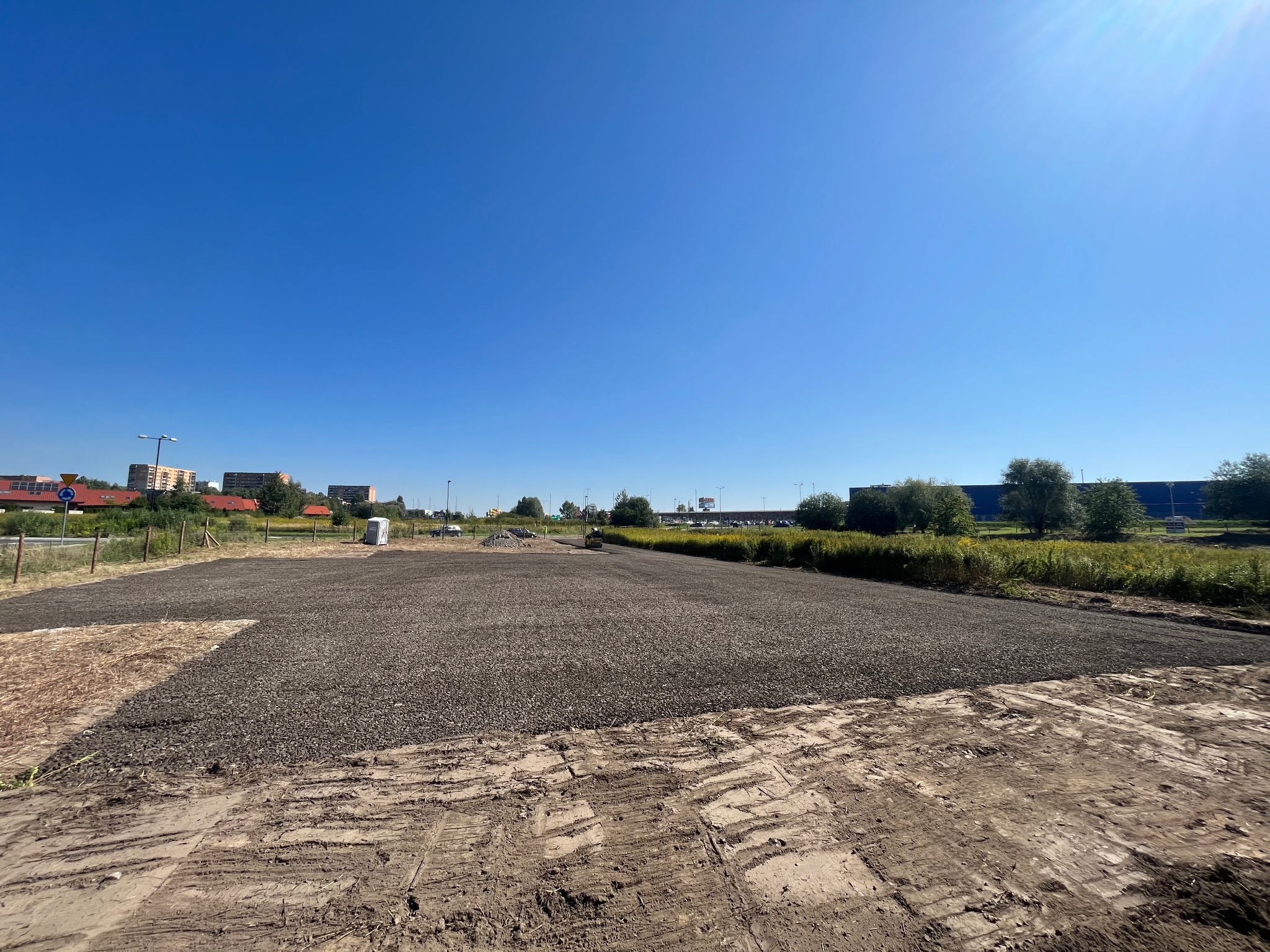 Teren po częściowym utwardzeniu drobnym kruszywem, widoczne ślady opon i budynki w tle, słoneczny dzień.