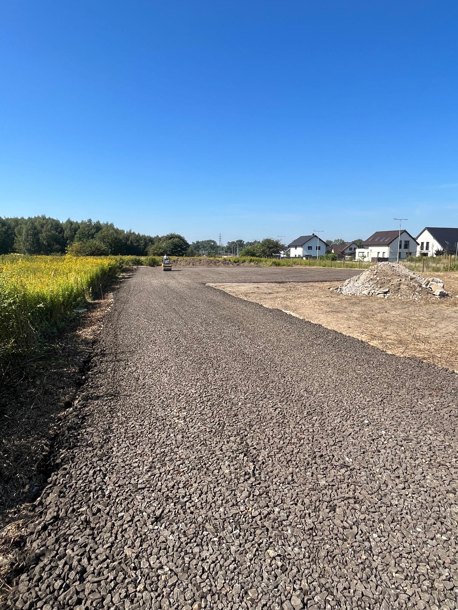 Utwardzona żwirowa droga dojazdowa na działce budowlanej, w tle domy jednorodzinne i zieleń. Walec drogowy w oddali. Słoneczny dzień.