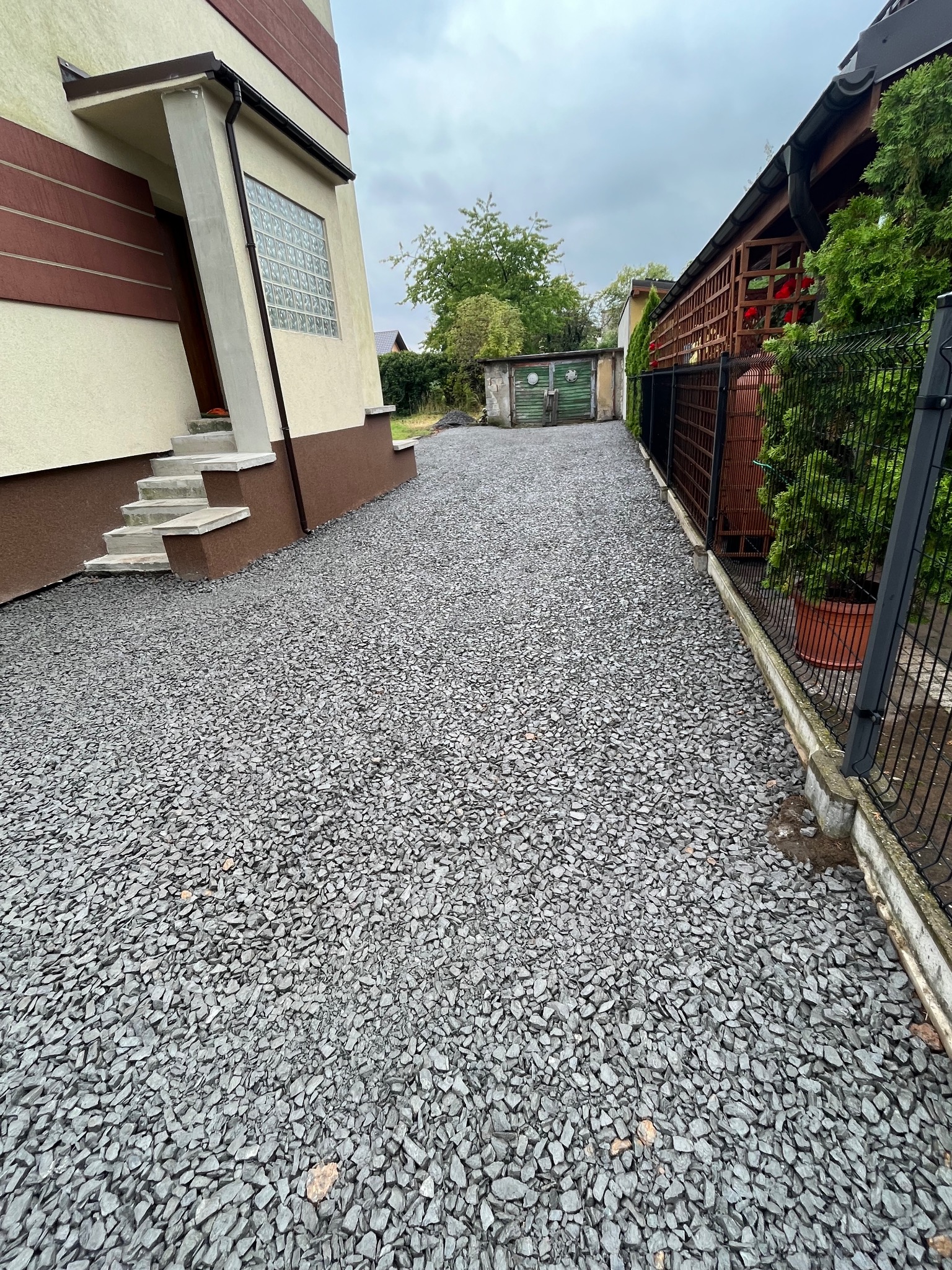 Szara, kamienna ścieżka prowadząca od schodów domu wzdłuż ogrodzenia. Widoczny budynek gospodarczy w tle. Ścieżka wykonana z drobnego, szarego gruzu.