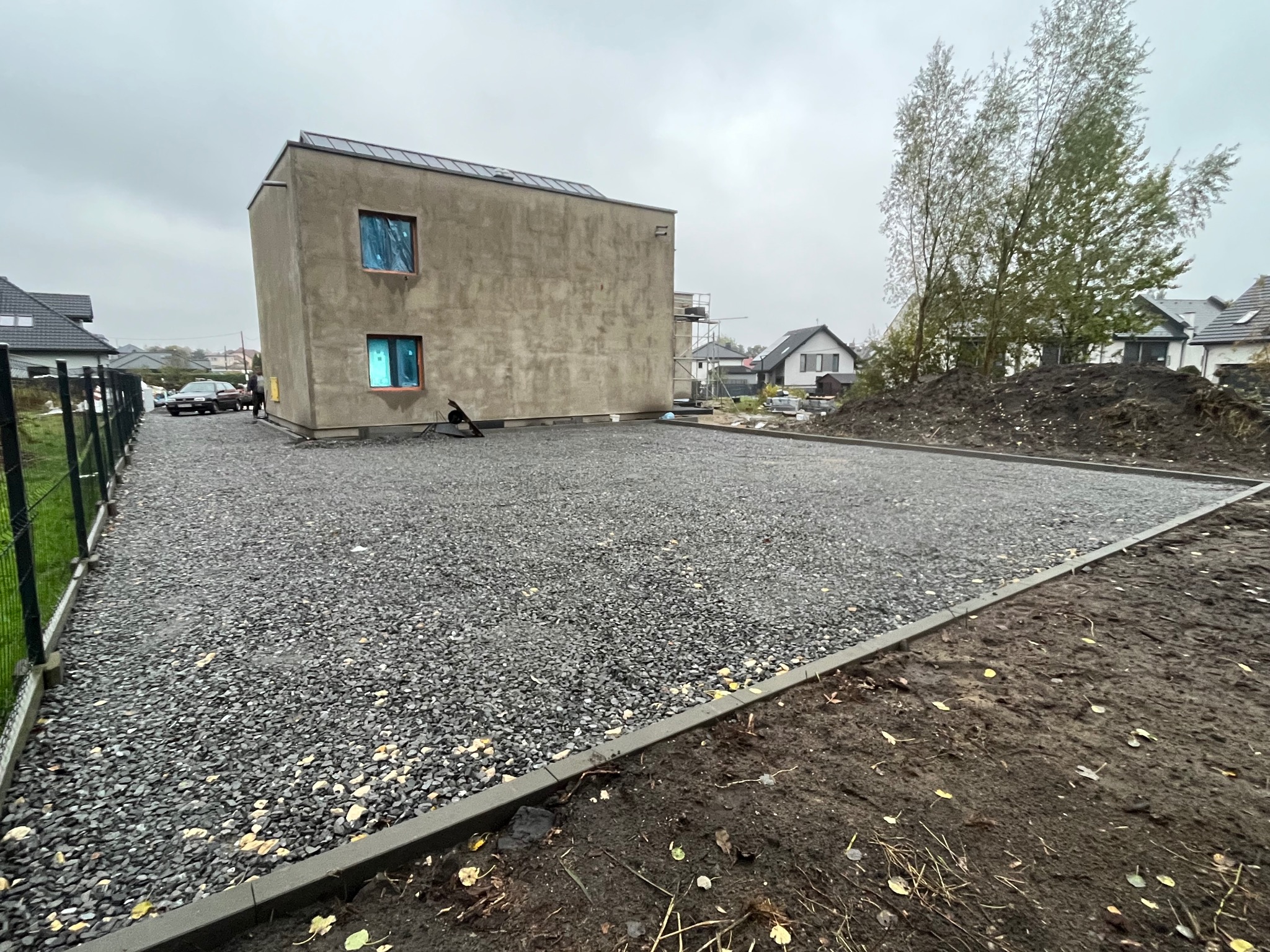 Utwardzony teren żwirem przy nowoczesnym domu z płaskim dachem, obrzeża z kostki. Widoczne drzewa i sąsiednie budynki w tle, pochmurne niebo.