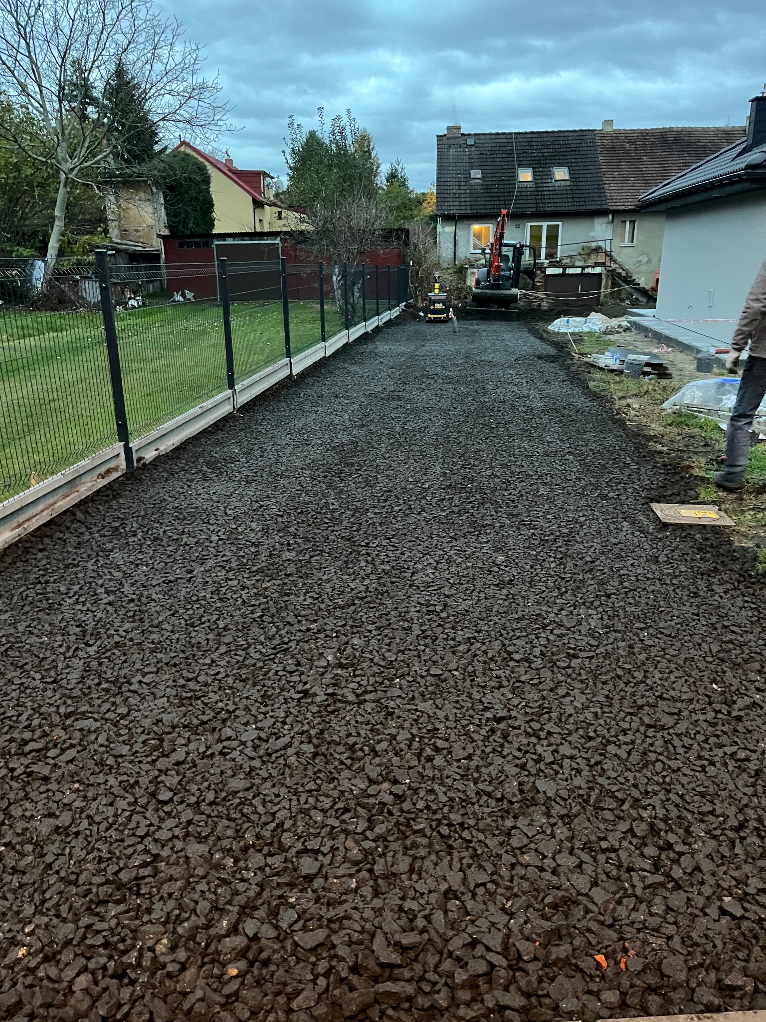 Świeżo położona, ciemna nawierzchnia z kruszywa na posesji, widoczny fragment ogrodzenia i budynki w tle. W oddali maszyna budowlana.