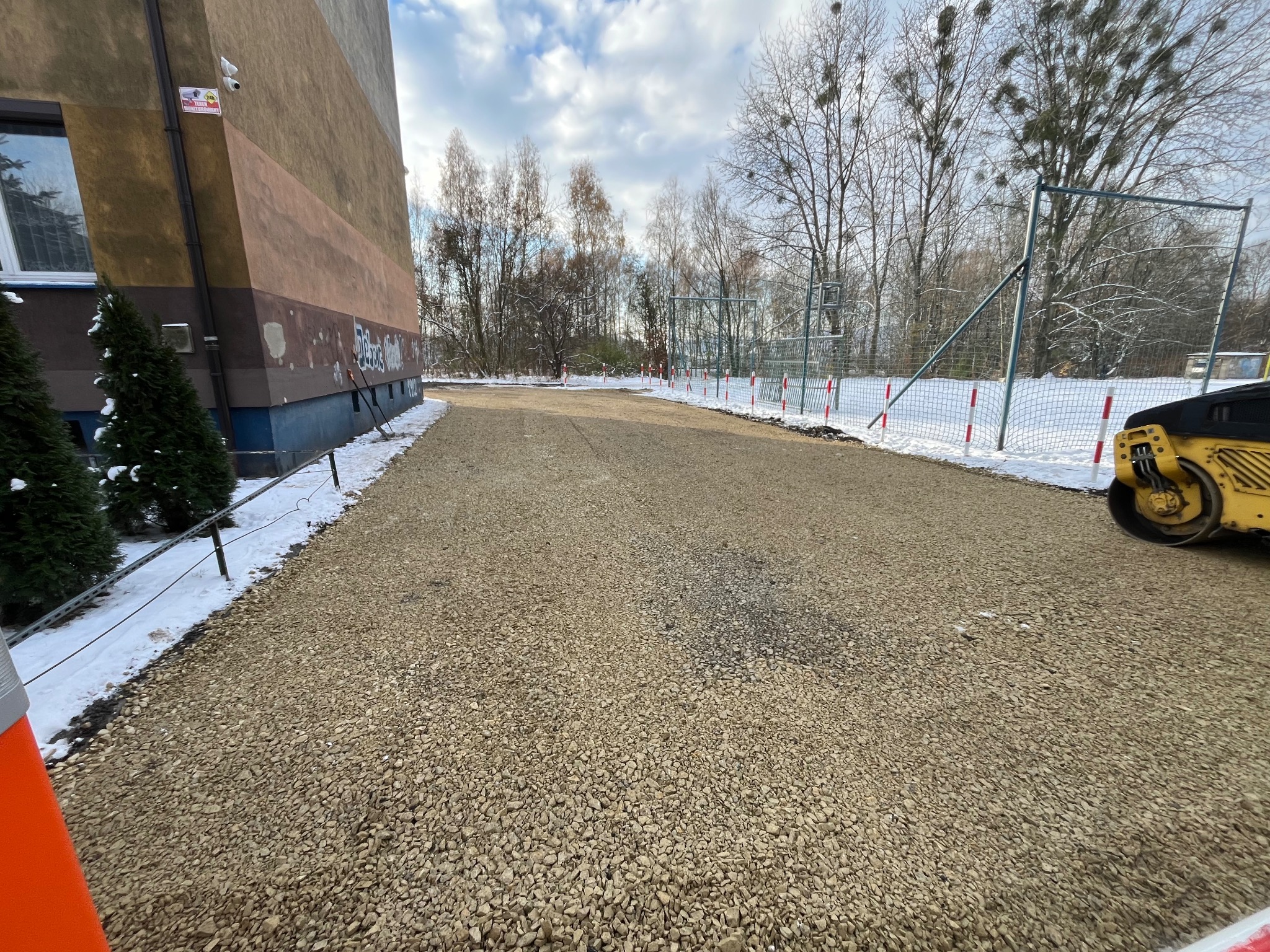Utwardzona droga żwirowa z widocznym walcem drogowym i elementami infrastruktury sportowej w tle, częściowo pokryta śniegiem.