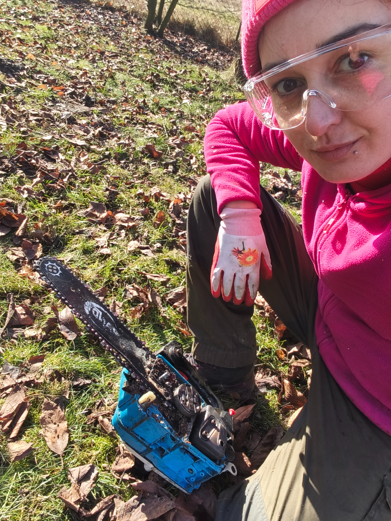 Kobieta w okularach ochronnych i różowej czapce siedzi na trawie z liśćmi obok niebieskiej piły łańcuchowej. Ubrana w rękawice robocze z kwiatowym wzorem.