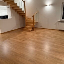 pro-desk-park - Jasna, drewniana podłoga i schody z przeszkloną balustradą w przestronnym wnętrzu. Minimalistyczne oświetlenie ścienne i okno z grzejnikiem. Widok na białą kuchnię w tle.