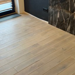 pro-desk-park - Jasna, drewniana podłoga ułożona w jodełkę, z widocznym fragmentem ściany w kolorze grafitowym i elementem dekoracyjnym imitującym marmur. Nowoczesne wykończenie wnętrza.