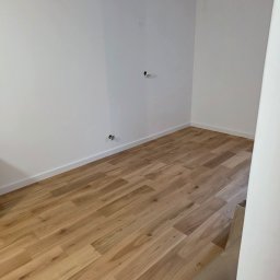 pro-desk-park - Jasne panele podłogowe ułożone w pustym, białym pomieszczeniu. Widoczne listwy przypodłogowe i wystające kable z sufitu. Fragment rolki wykładziny w rogu.