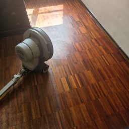 pro-desk-park - Cyklinowanie parkietu w trakcie realizacji. Widoczna maszyna do cyklinowania na błyszczącej, odnowionej drewnianej podłodze. Parkiet ułożony w regularny wzór.