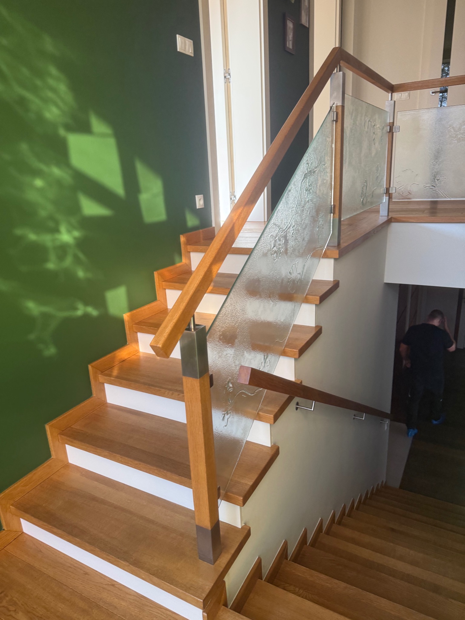 Drewniane schody z przeszkloną balustradą, oświetlone naturalnym światłem, z widocznym fragmentem zielonej ściany i postacią w tle. Detal wykończenia wnętrza.