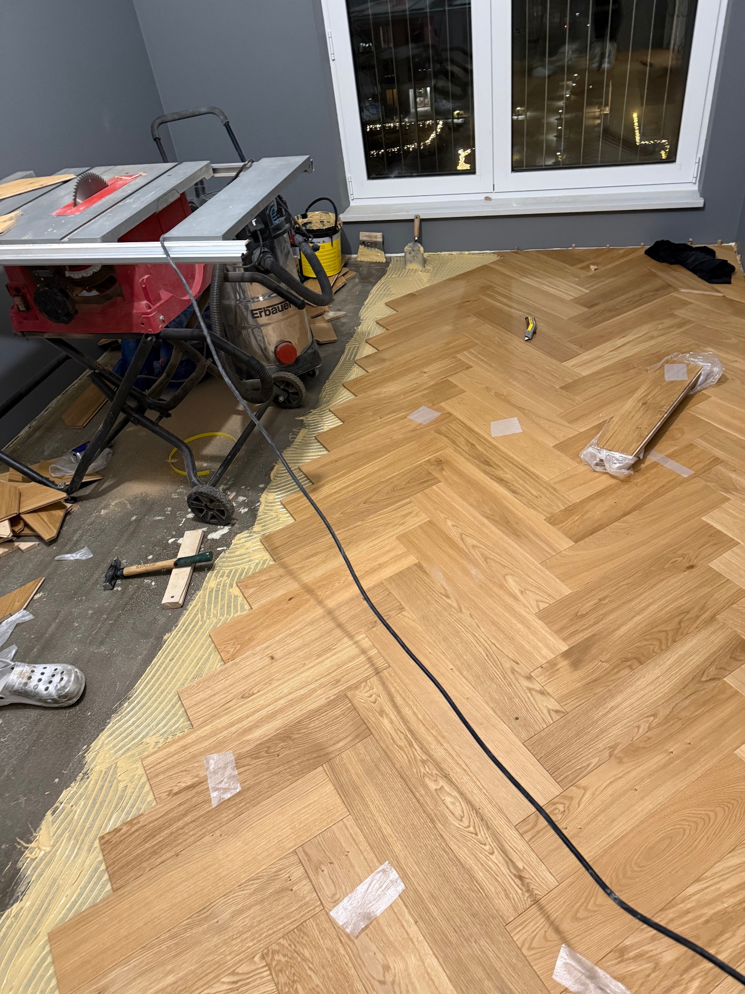 Układanie parkietu w jodełkę: widoczny fragment ułożonej podłogi, narzędzia (piła, odkurzacz, młotek) i klej. Praca w toku, z widokiem na okno i nocne miasto.