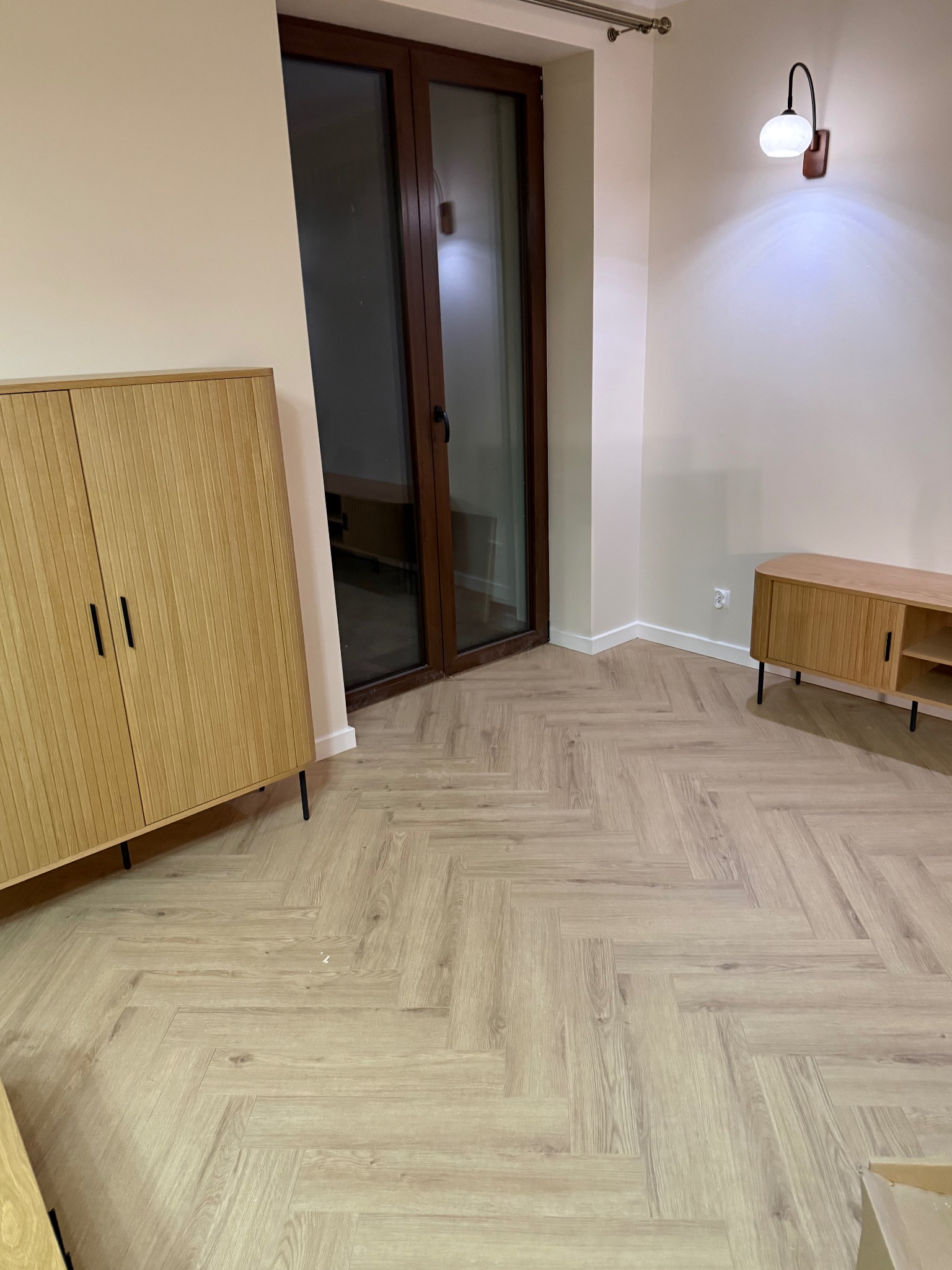 Jasna podłoga ułożona w jodełkę francuską w nowocześnie urządzonym pokoju z drewnianymi meblami i oknem balkonowym. Minimalistyczne oświetlenie.