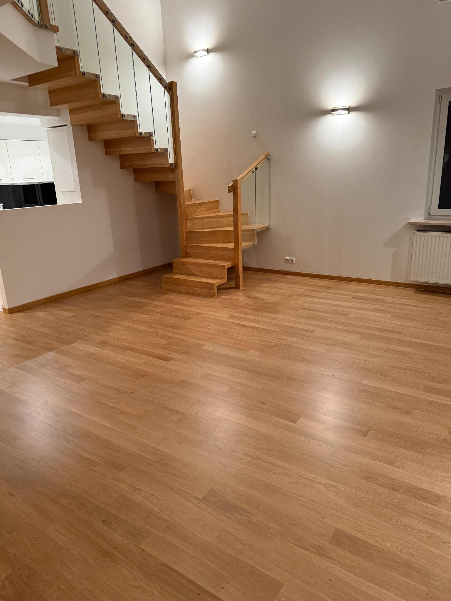 Jasna, drewniana podłoga i schody z przeszkloną balustradą w przestronnym wnętrzu. Minimalistyczne oświetlenie ścienne i okno z grzejnikiem. Widok na białą kuchnię w tle.