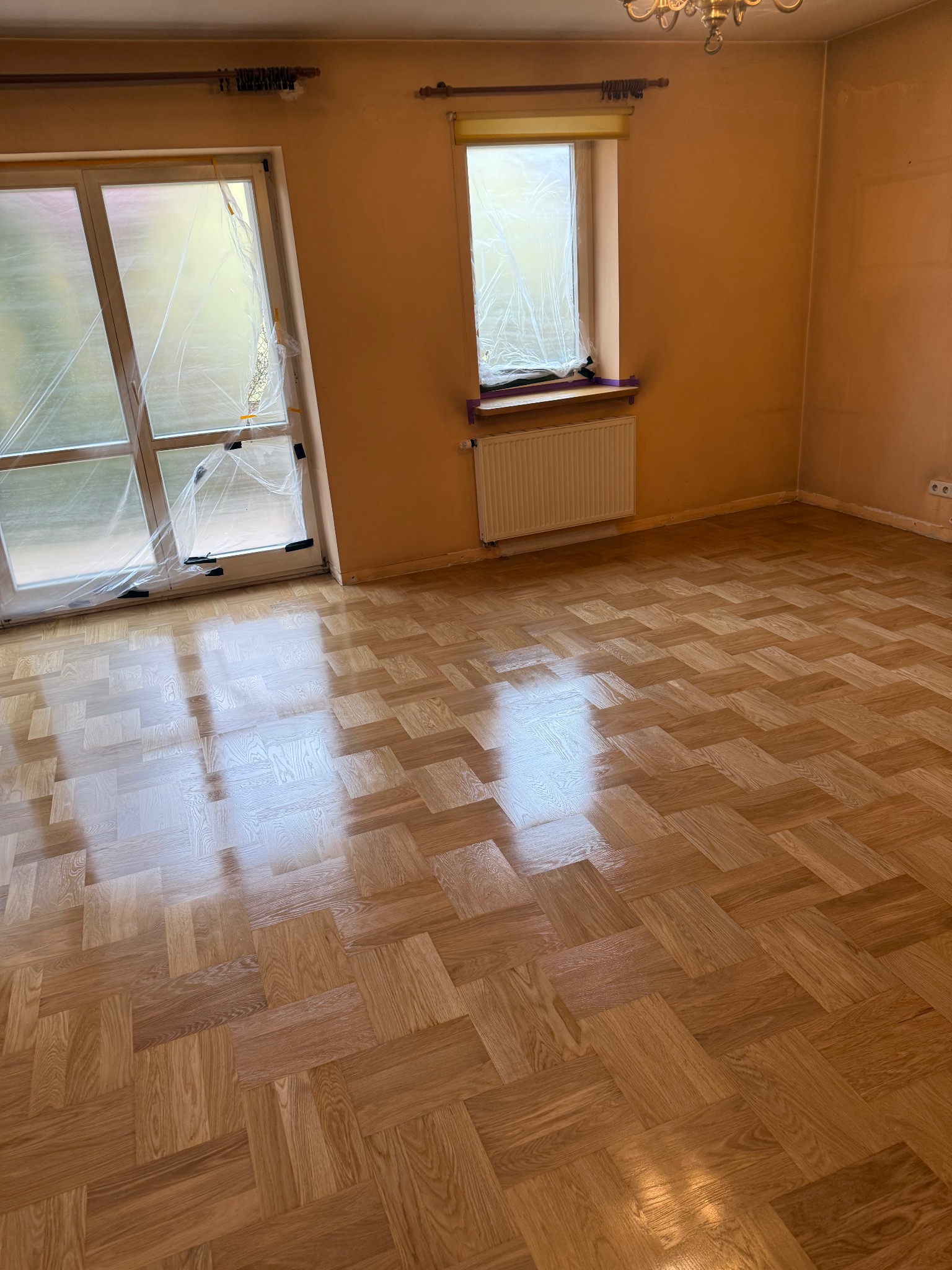 Błyszcząca, nowa podłoga parkietowa ułożona w jodełkę w pustym pokoju. Okna i grzejnik zabezpieczone folią malarską. Ściany w odcieniach beżu.