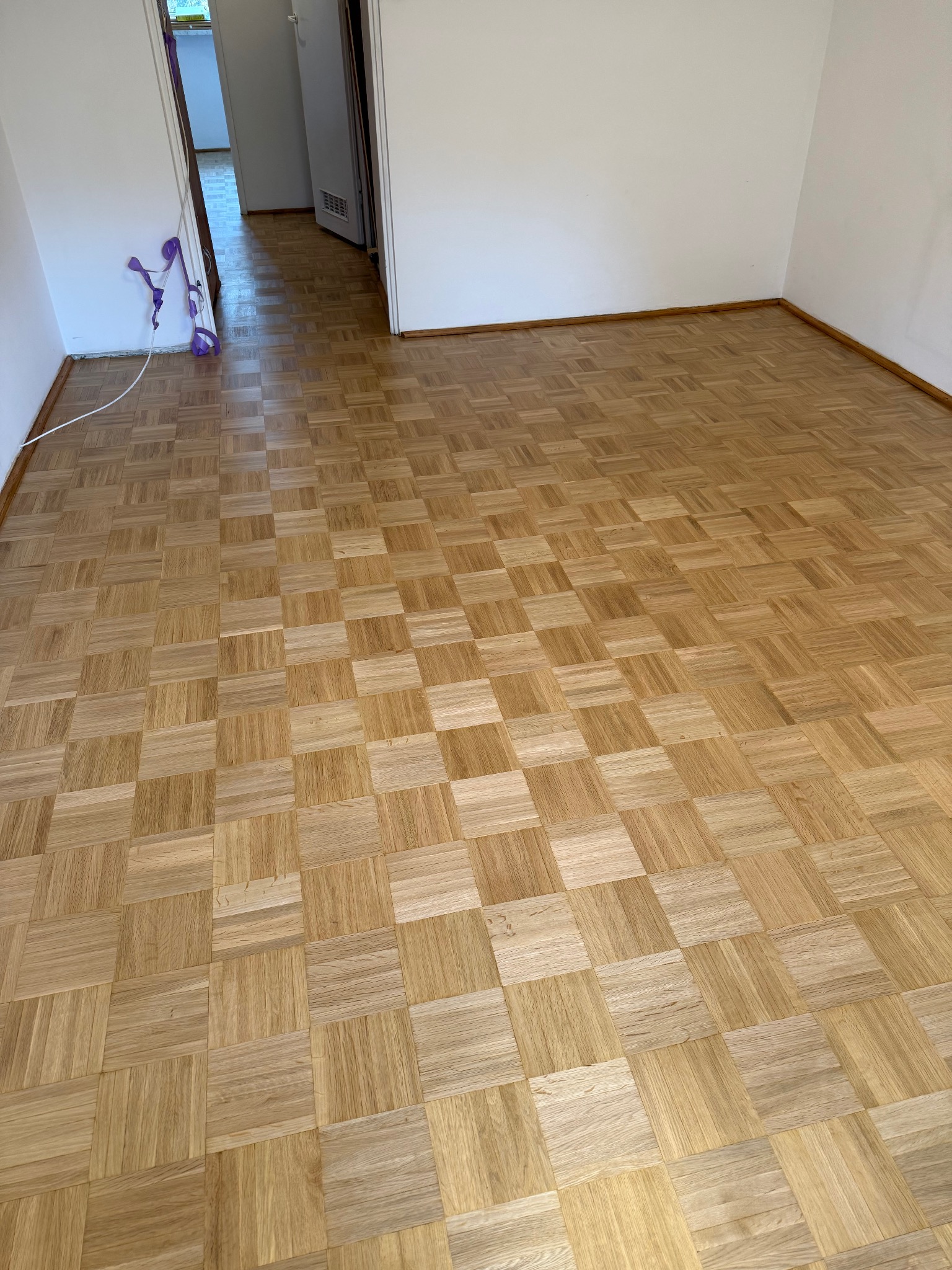 Parkiet z jasnego drewna ułożony w kwadraty w pustym pokoju z białymi ścianami. Widoczne listwy przypodłogowe i otwarte drzwi do kolejnego pomieszczenia.