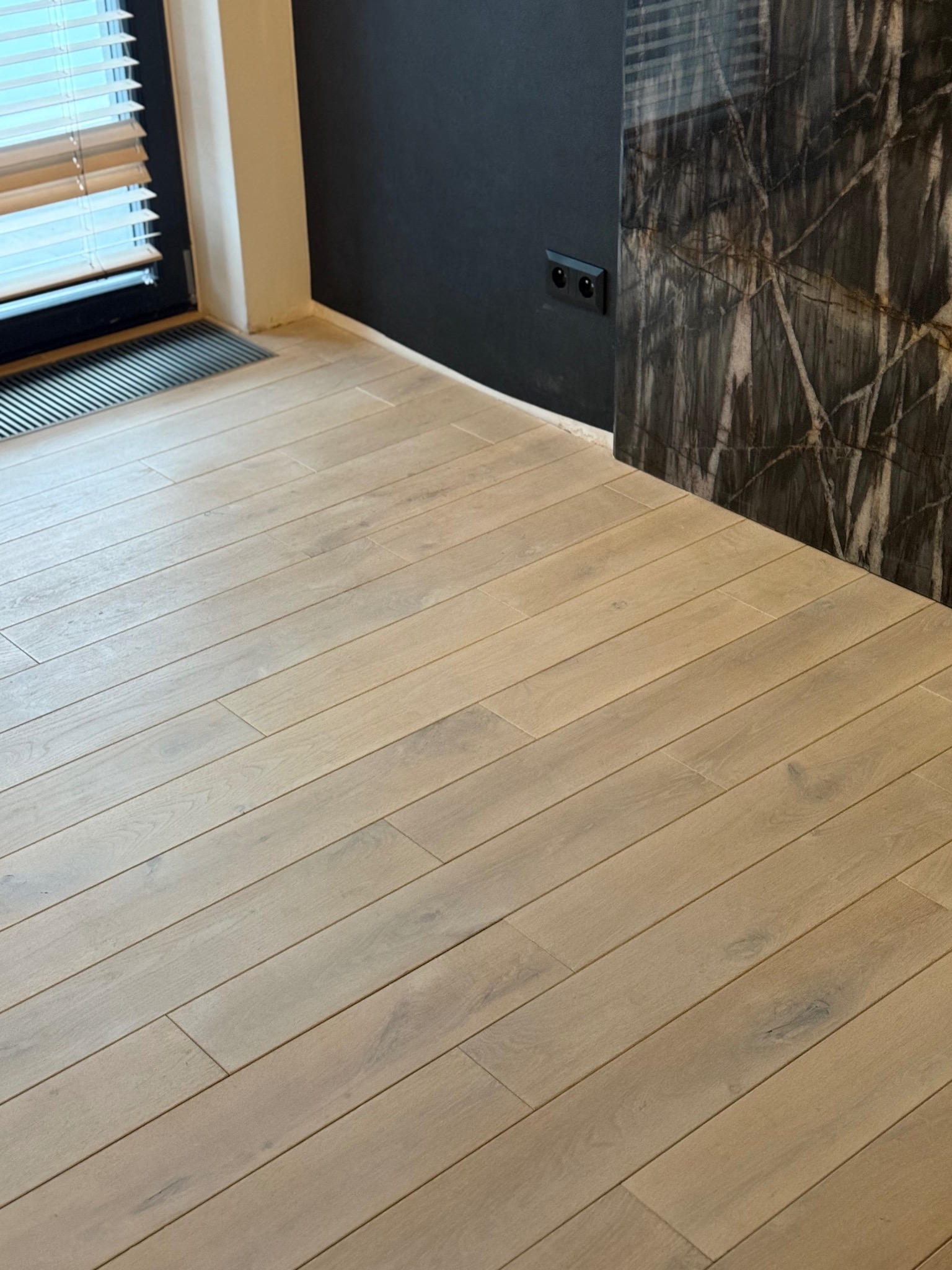 Jasna, drewniana podłoga ułożona w jodełkę, z widocznym fragmentem ściany w kolorze grafitowym i elementem dekoracyjnym imitującym marmur. Nowoczesne wykończenie wnętrza.