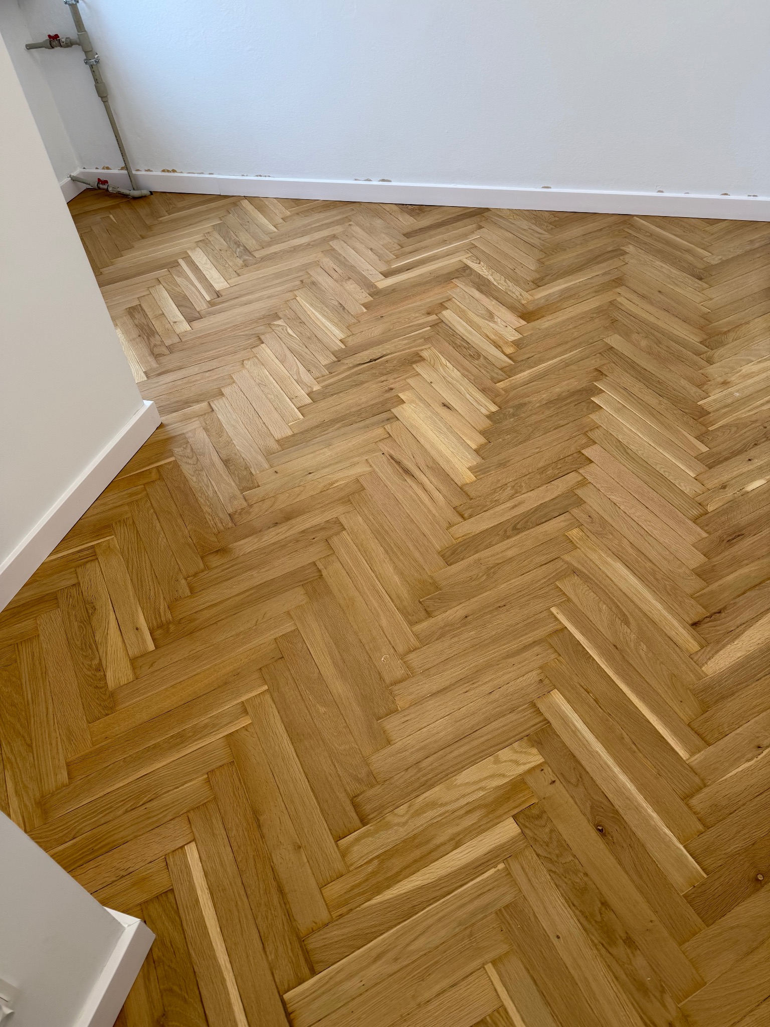 Ułożony parkiet w jodełkę, jasne drewno, białe listwy przypodłogowe, widok z góry, fragment instalacji przy ścianie. Naturalne światło.