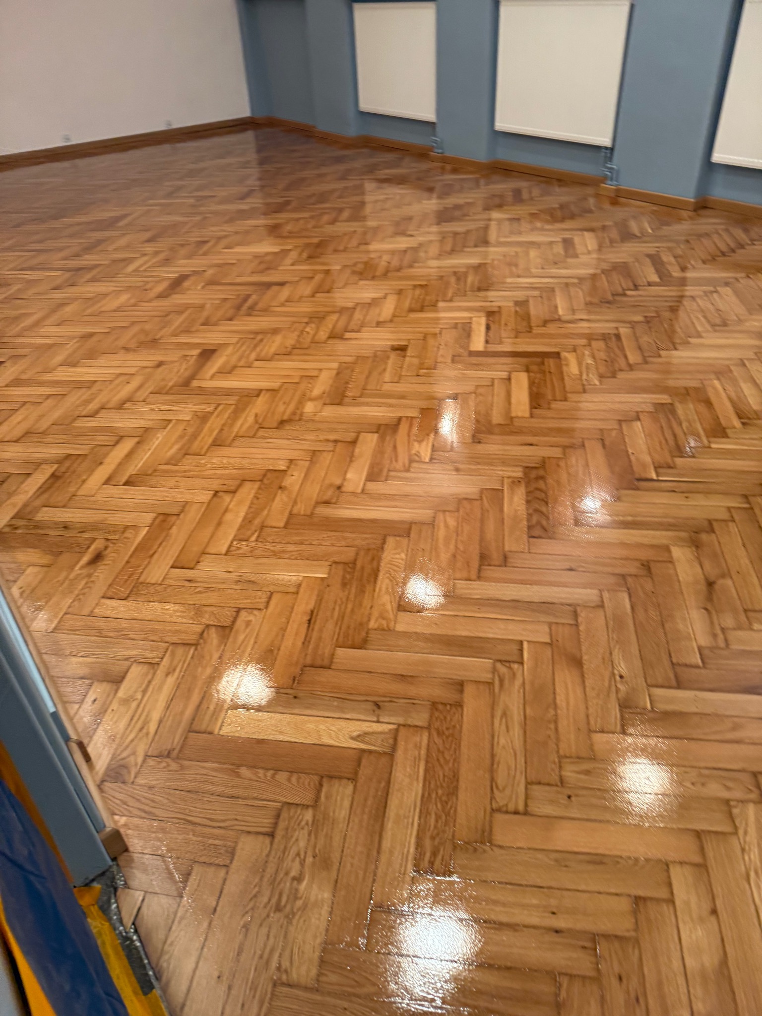 Błyszcząca, świeżo polakierowana podłoga parkietowa ułożona we wzór jodełki francuskiej, widok z niskiej perspektywy, fragment osłony zabezpieczającej w lewym dolnym rogu.