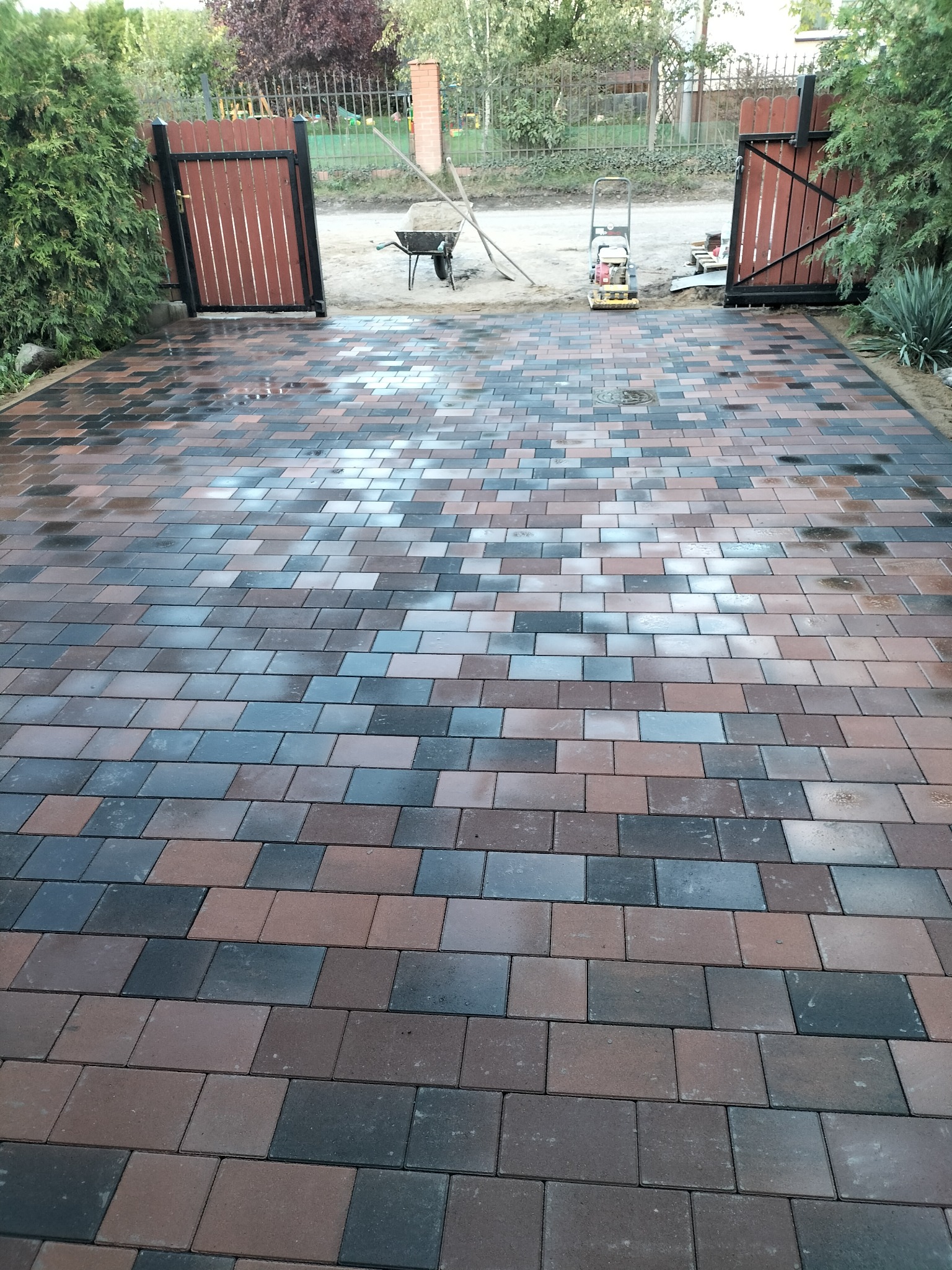 Ułożona kostka brukowa w odcieniach brązu i szarości, prowadząca do otwartej, drewnianej bramy. W tle widoczny sprzęt budowlany i zieleń.