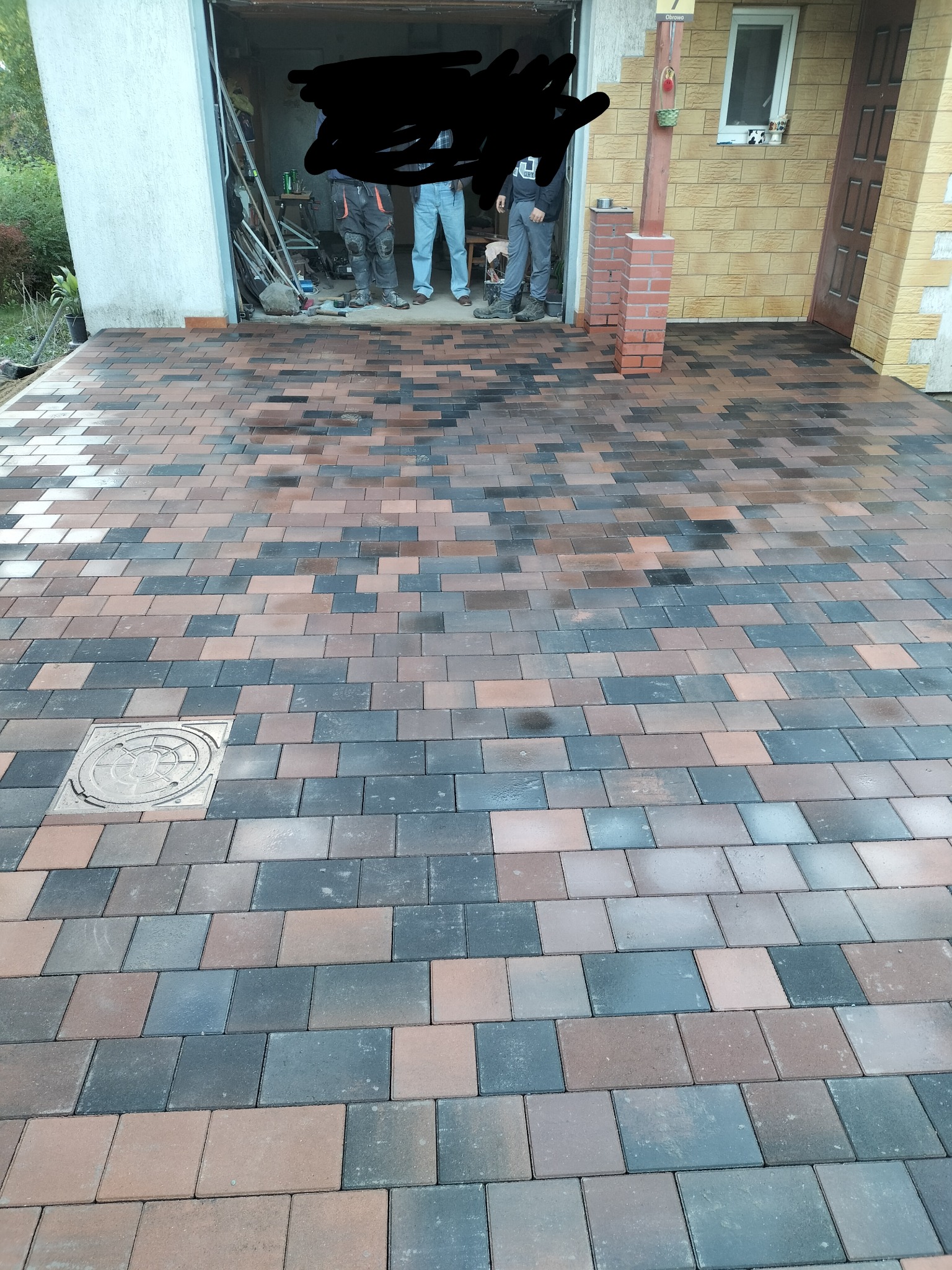 Ułożona kostka brukowa w odcieniach brązu i szarości przed garażem, z widoczną studzienką kanalizacyjną. W tle osoby i narzędzia w otwartym garażu.