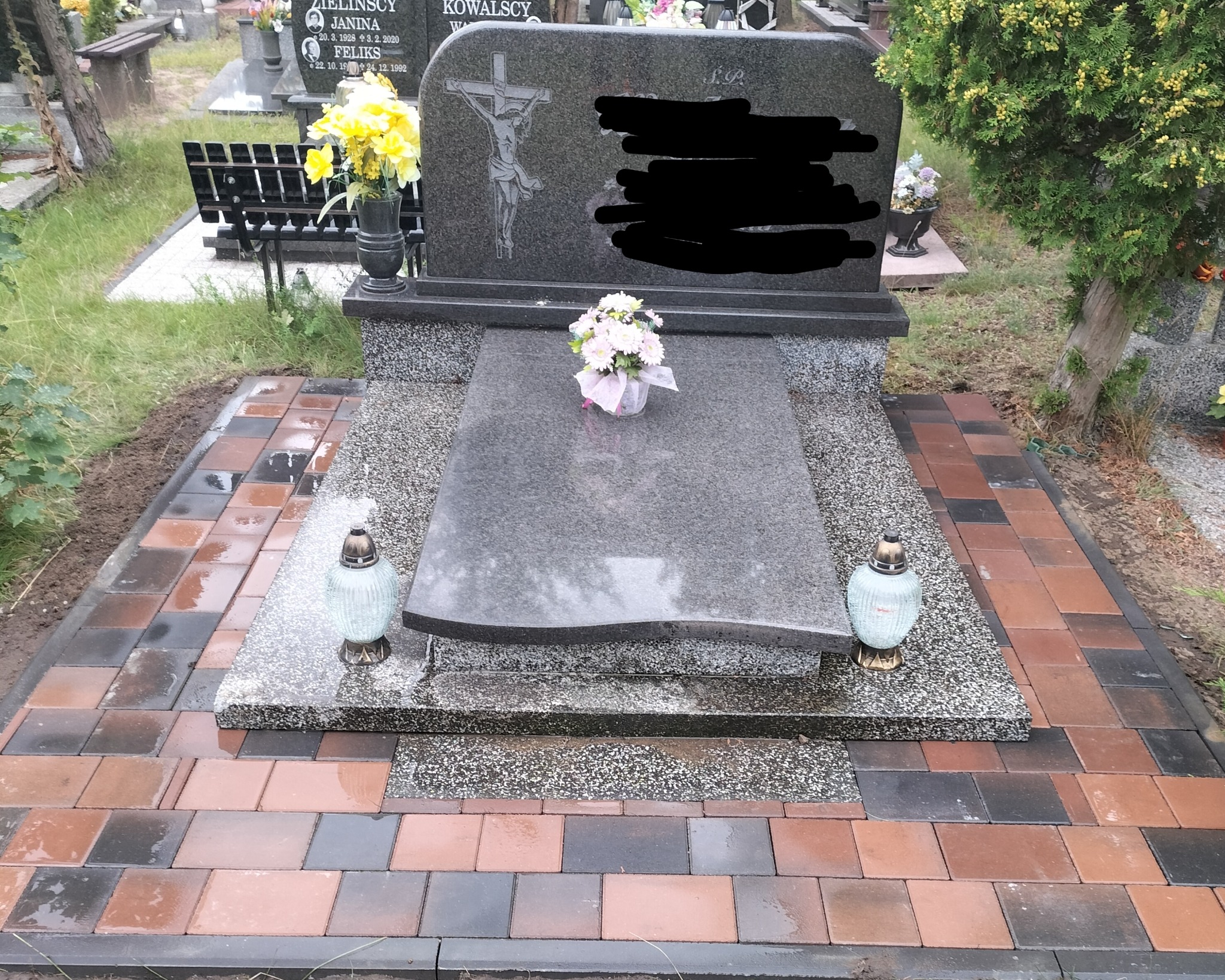 Granitowy nagrobek z krzyżem i kwiatami, otoczony nową, kolorową kostką brukową. Widoczne lampiony i fragmenty innych nagrobków na cmentarzu.