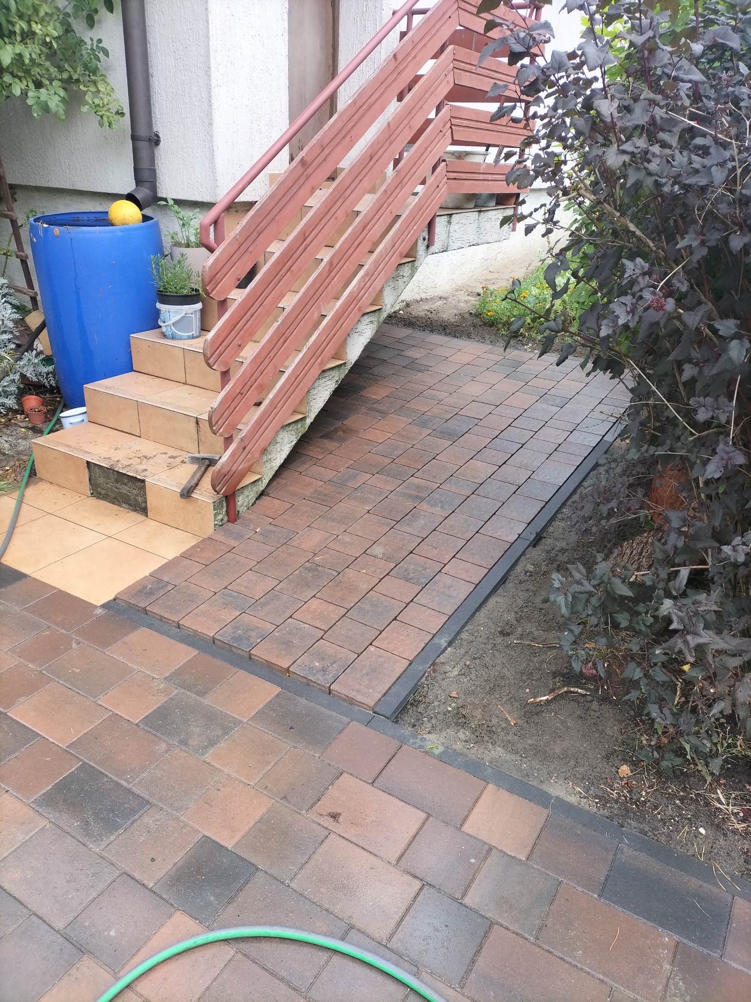 Ułożona kostka brukowa prowadząca do schodów wejściowych, z widocznym fragmentem trawnika i roślinności. Brązowo-szara kostka, schludne wykończenie.