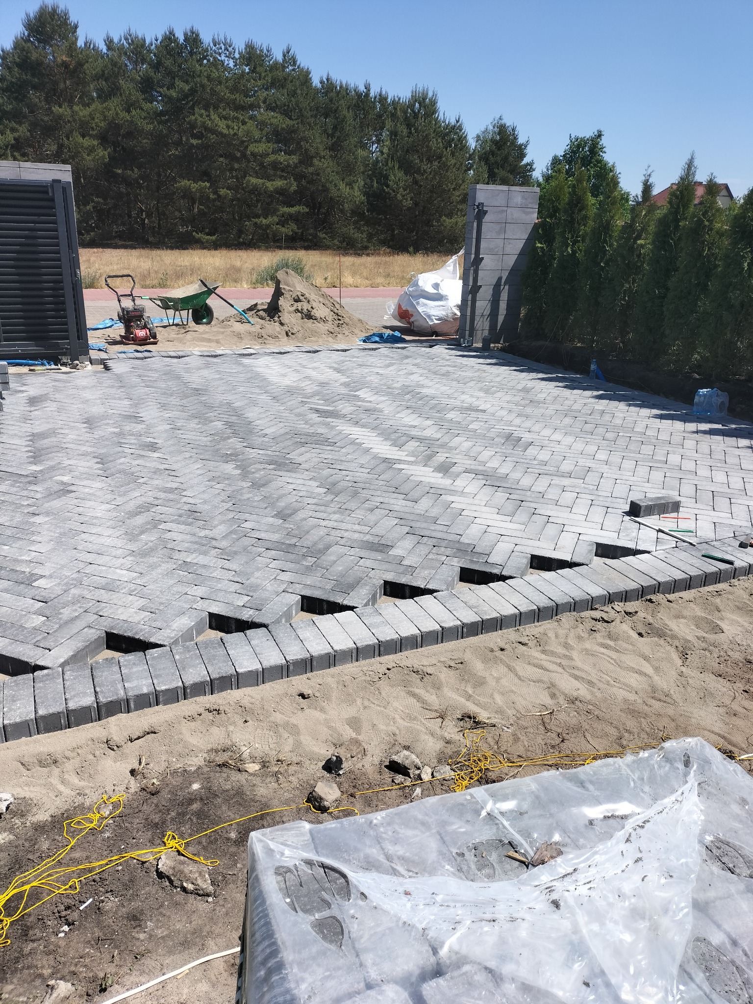 Układanie szarej kostki brukowej w jodełkę na podjeździe. Widoczne niedokończone fragmenty, piasek, narzędzia i brama w tle. Słoneczny dzień.