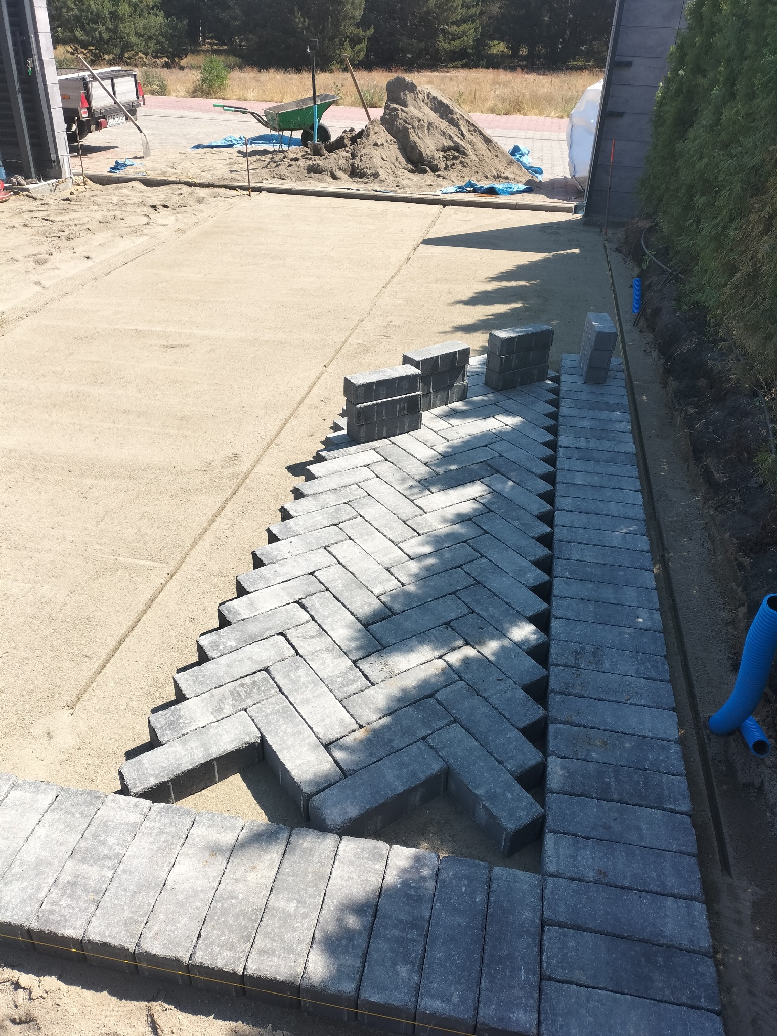 Fragment układania szarej kostki brukowej w jodełkę na podsypce piaskowej. Widoczne narzędzia i materiały budowlane w tle. Częściowo ułożone obrzeże.