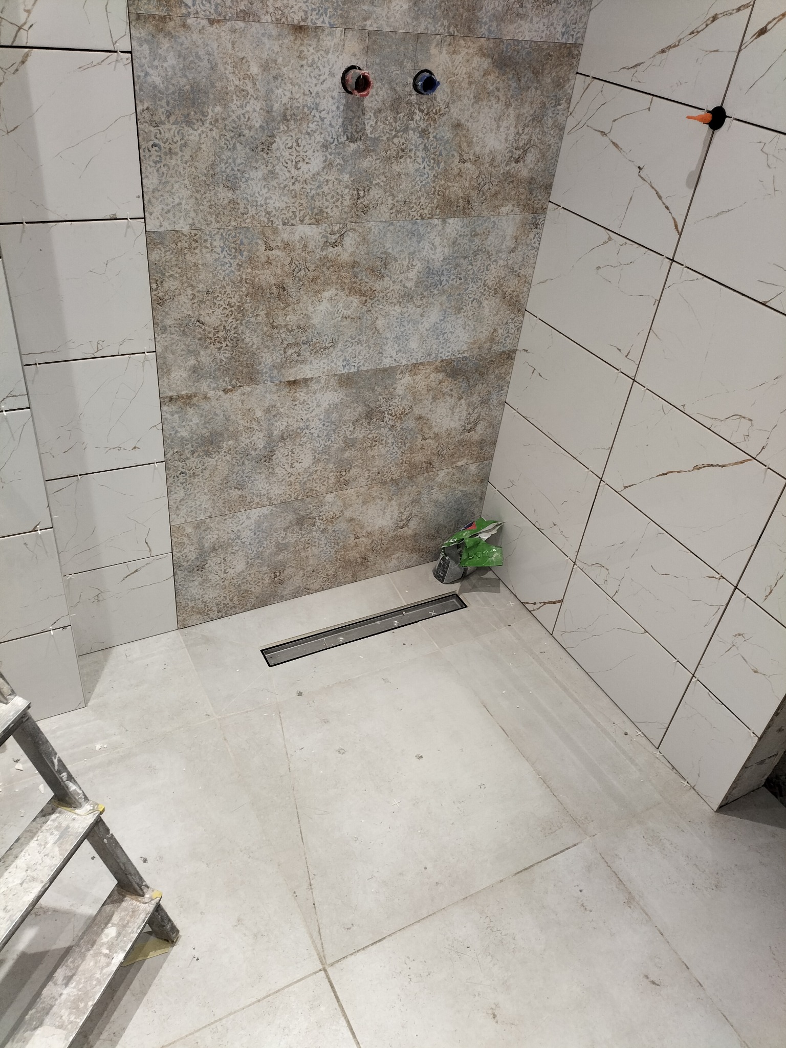 Wnętrze łazienki w trakcie remontu: ułożone płytki ścienne w dwóch wzorach, odpływ liniowy w podłodze, drabina malarska w rogu. Widoczne krzyżyki dystansowe.