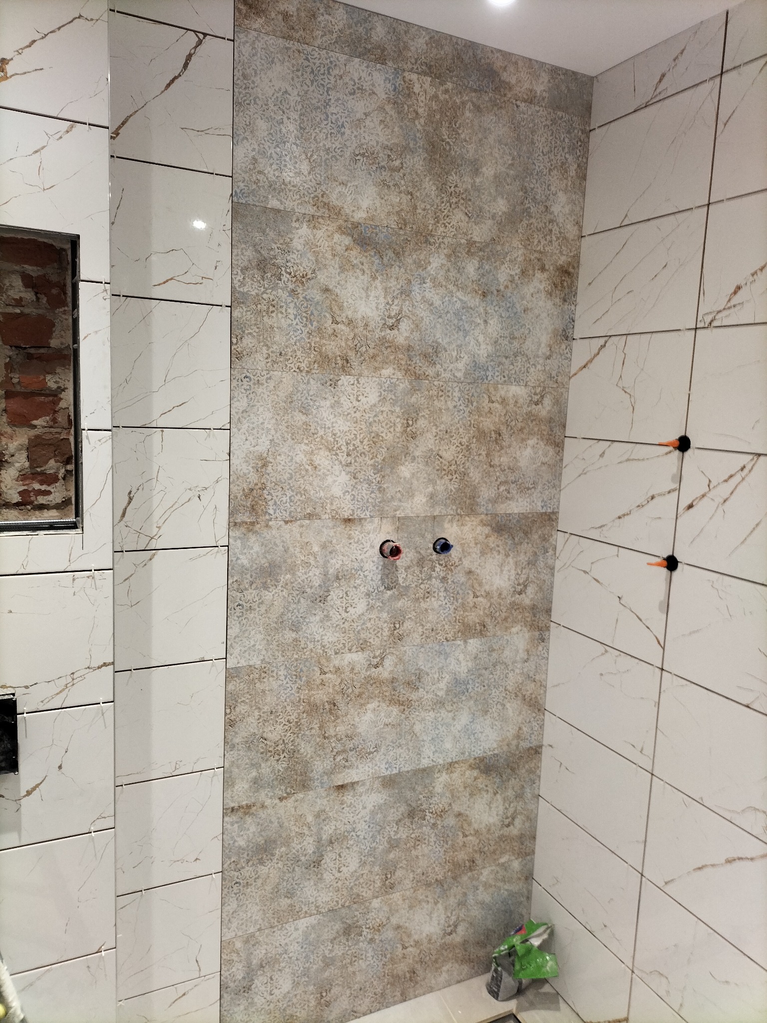 Wykończona ściana w łazience z białymi płytkami imitującymi marmur i dekoracyjnymi płytkami w odcieniach beżu i szarości. Widoczne otwory na baterie i krzyżyki dystansowe.