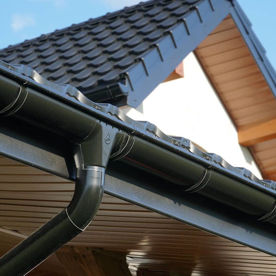 Czarne, metalowe rynny zamontowane na dachu z ciemnej dachówki, widoczne na tle błękitnego nieba i fragmentu białej ściany budynku. Ujęcie z dołu.