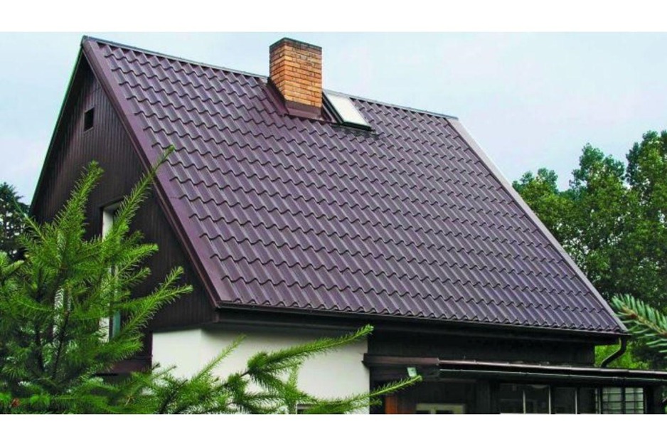 Dom z ciemnobrązowym dachem z blachodachówki, ceglanym kominem i oknem dachowym. Widok z dołu, zieleń w tle.