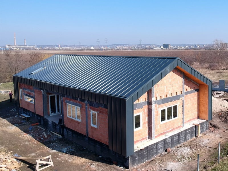 Nowoczesny dom w trakcie budowy z ciemnoszarym dachem z blachy na rąbek stojący i częściowo wykończoną elewacją z cegły. Widok z lotu ptaka.