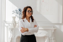 Uśmiechnięta kobieta w okularach, białej koszuli i czarnych spodniach stoi na tle białego kominka i dekoracji, prezentując profesjonalny wizerunek.