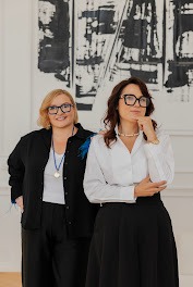 Dwie uśmiechnięte kobiety w okularach, elegancko ubrane w czarno-białe stroje, stoją na tle abstrakcyjnego obrazu w biurze. Jedna z piórami na ramieniu.