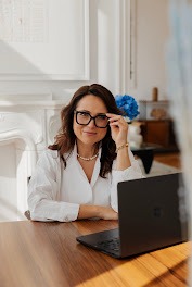 Uśmiechnięta kobieta w okularach siedzi przy biurku z laptopem w jasnym biurze. W tle widoczny kominek i dekoracje.