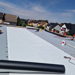 Fach plaski dach - Biała termoizolacja na szarym dachu płaskim w trakcie montażu, widoczne narzędzia i rolka papy. W tle zabudowa jednorodzinna i błękitne niebo.