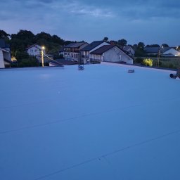 Fach plaski dach - Nowy, płaski dach pokryty jasną membraną na tle zabudowań mieszkalnych o zmierzchu. Widok z góry ukazuje gładką powierzchnię i detale wykończenia.